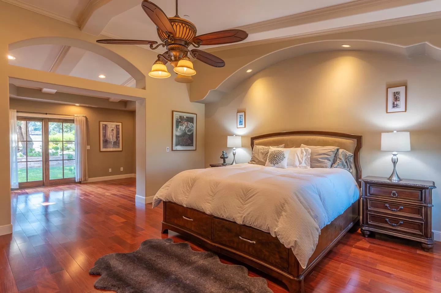 Spacious bedroom with wooden floors, a large bed with white bedding, and cozy lighting. Large glass doors open to a garden, beautifully showcasing the quality you can expect from a general contractor Pensacola trusts for home construction.