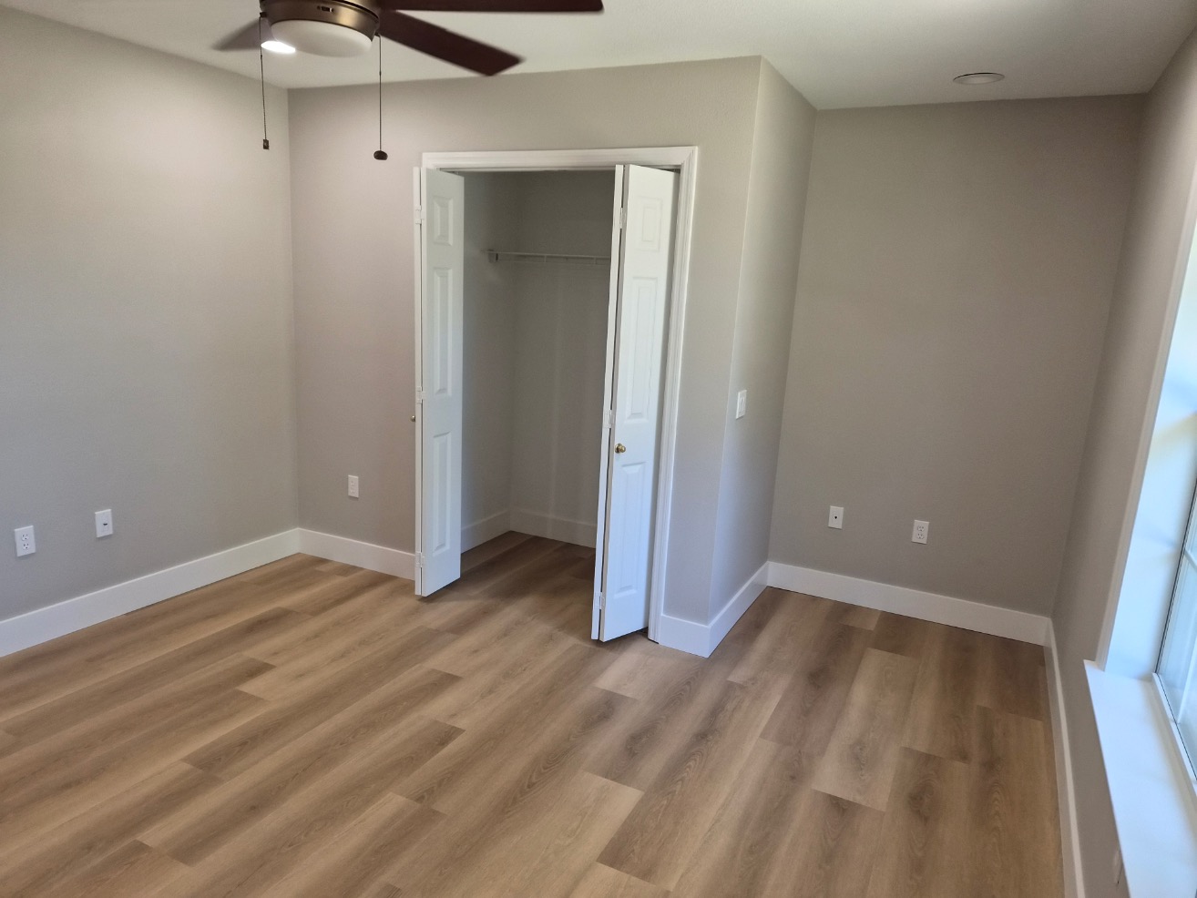 A bright, empty room with light wood flooring, beige walls, a ceiling fan, double-door closet, electrical outlets, and a large window letting in natural light—perfect for your next Pensacola remodeling or home construction project.