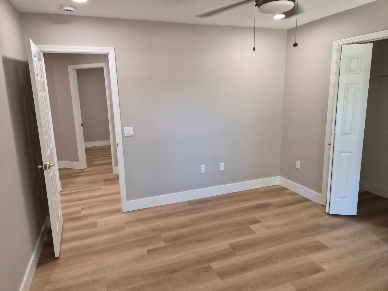 Empty room with light wood flooring, light gray walls, white trim, open closet door, ceiling fan, and two open doors leading to adjacent rooms—ideal for a Pensacola remodeling project or a home construction upgrade. No furniture or decorations are present.