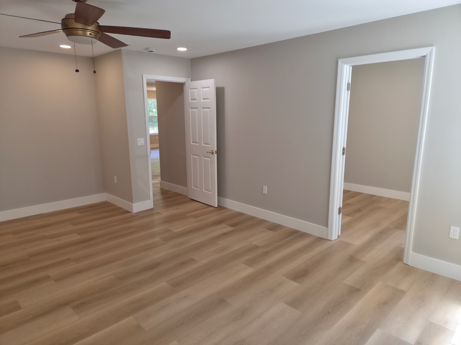 Empty room with light wood flooring, beige walls, white trim, a ceiling fan, recessed lighting, and two open doorways leading to adjoining rooms—perfect for your next Pensacola remodeling or home construction project. Natural light enters through a window.