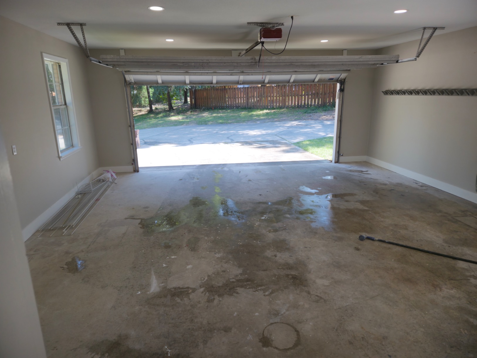A mostly empty garage with a partially open door, some water and stains on the concrete floor, a window on the left, shelving on the right wall—ideal for home construction storage or as a staging area for your next Pensacola remodeling project.