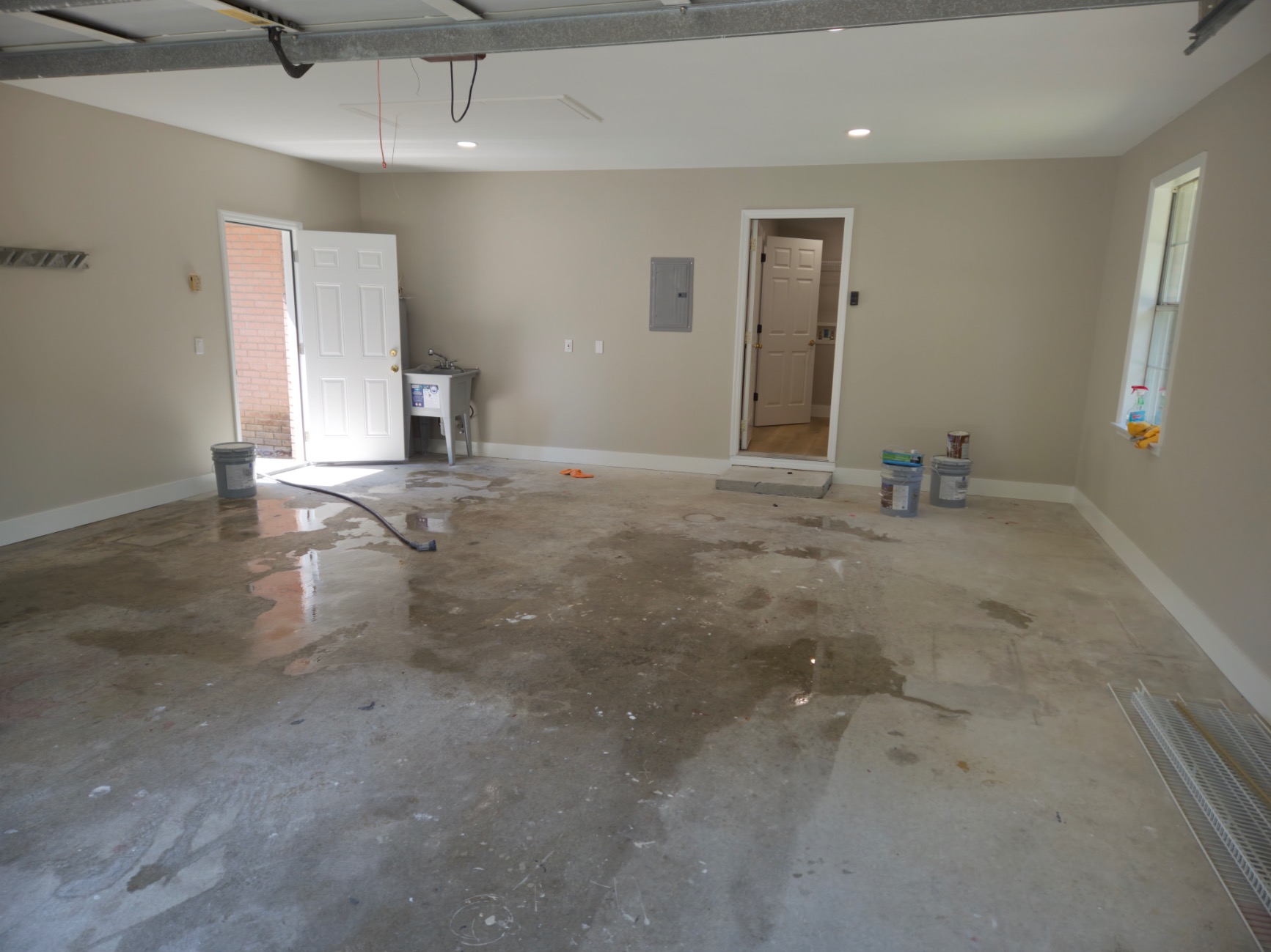 A mostly empty garage with a wet, stained concrete floor, some paint cans and cleaning supplies against the walls—ideal for storing tools during your Pensacola remodeling or home construction project—with doors to both the outside and inside the house.