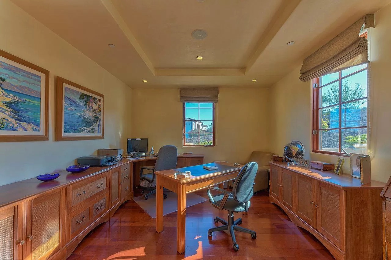 A home office with a wooden desk and chair in the center, side desks with a computer, globe, and framed art on the walls. Sunlight streams through a window showing trees and mountains—ideal inspiration for your next Pensacola remodeling project.