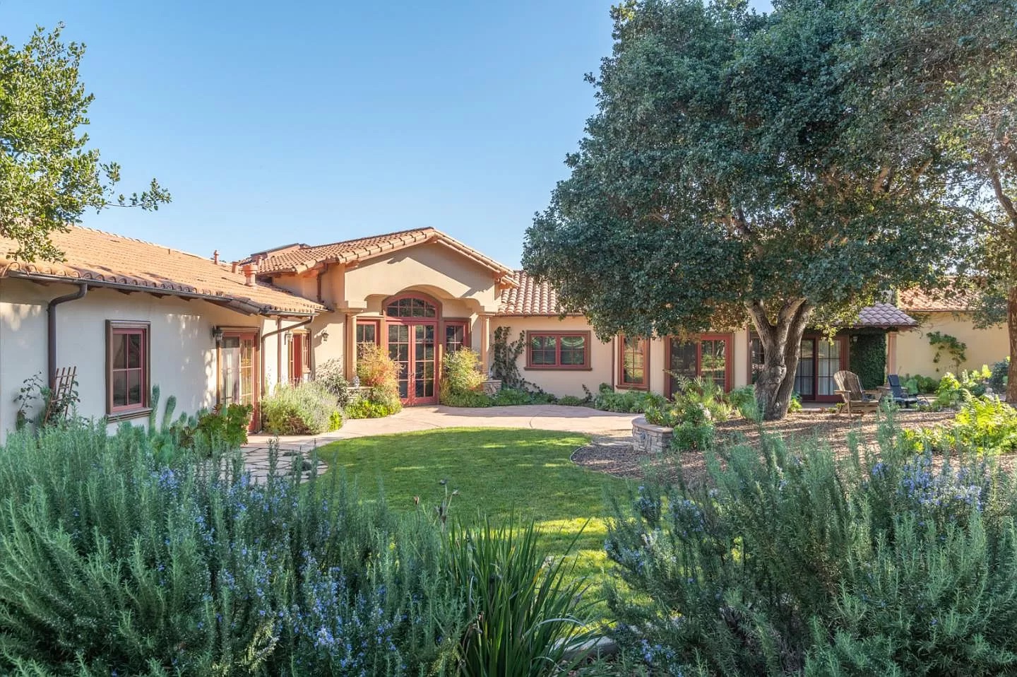 Single-story Mediterranean-style house with a red-tiled roof, large windows, and arched entryway, surrounded by lush landscaping—a perfect inspiration for your next Pensacola remodeling or bathroom remodel project under a clear blue sky.