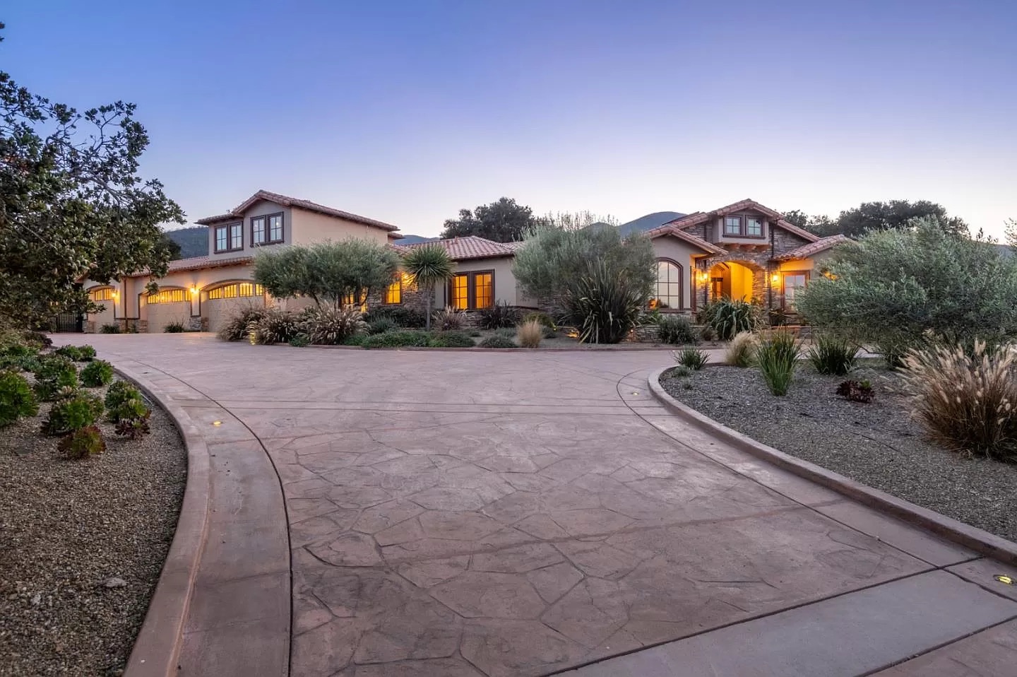 A large, elegant house with warm exterior lights, multiple peaked roofs, and dormer windows sits beside a wide, curved driveway bordered by desert landscaping at dusk—expertly crafted by a leading general contractor in Pensacola.