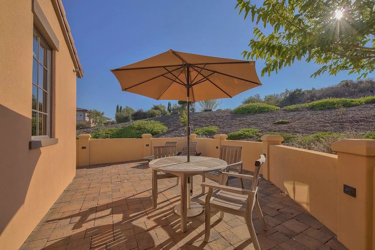 A sunny patio with a round table, four wooden chairs, and a large umbrella. The area is paved with bricks by an expert general contractor Pensacola trusts, surrounded by a low yellow wall with trees, bushes, and a hillside in the background.