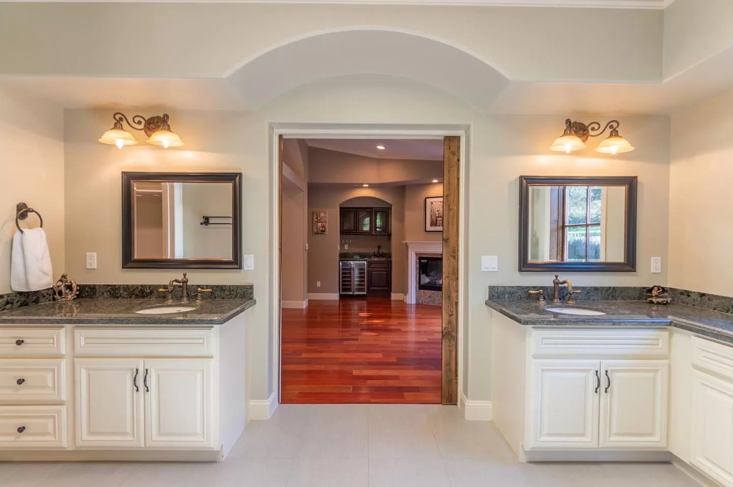 Spacious bathroom with two vanities, each with a mirror and lighting, cream cabinets, and dark countertops. An open doorway reveals wood floors and decor—perfectly showcasing quality Pensacola remodeling by a trusted general contractor.
