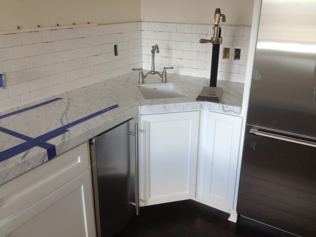 A modern kitchen corner with white cabinets, marble countertop, small sink with double-handle faucet, subway tile backsplash, and stainless steel appliances—perfect inspiration for your Pensacola remodeling or kitchen remodel project. Blue tape marks are visible on the counter.