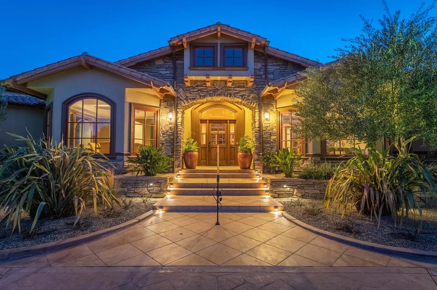 A warmly lit house with stone and stucco exterior, large windows, and a wooden double door. Steps with lights lead to the entrance, surrounded by landscaped plants—showcasing top-tier home construction by your general contractor in Pensacola.