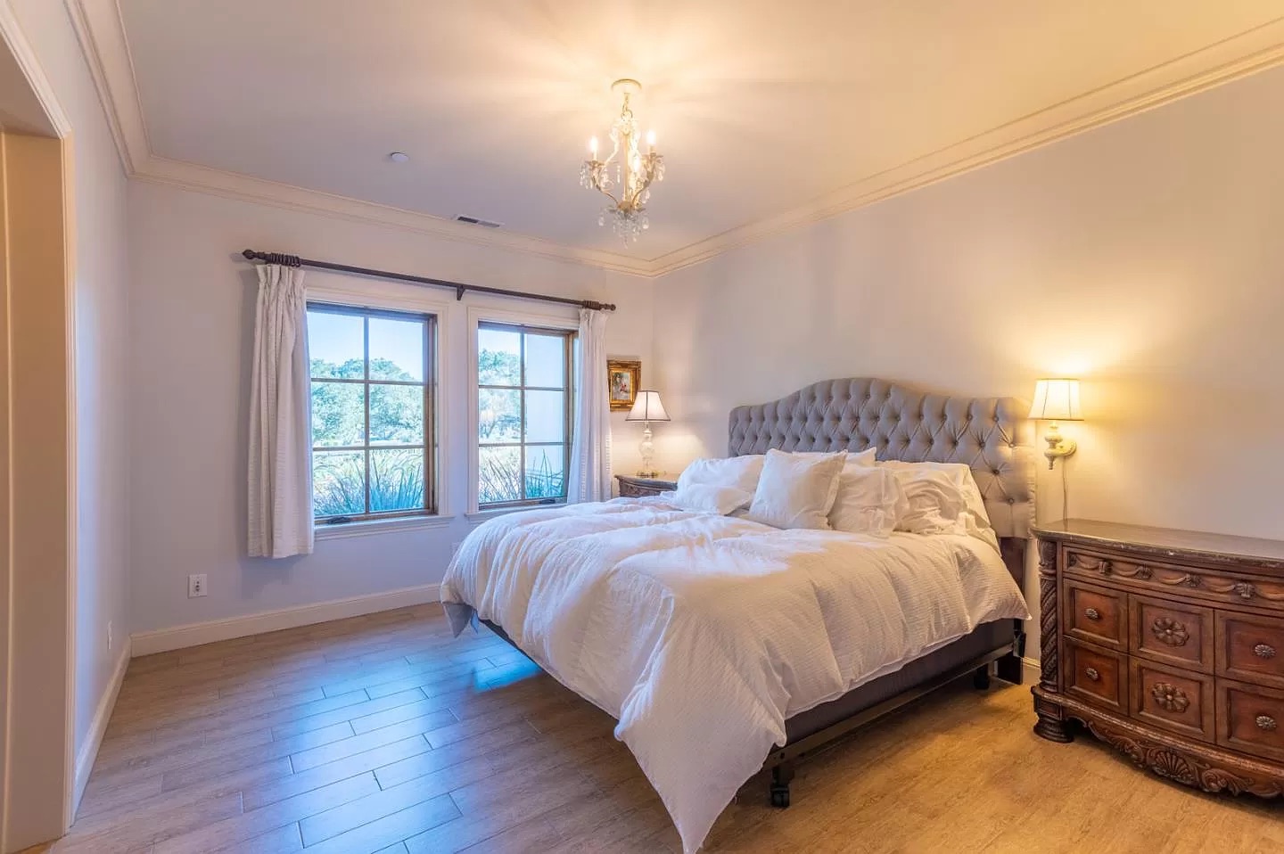 A bright bedroom in a Pensacola remodeling project features a large tufted bed, white bedding, two bedside lamps, a wooden dresser, chandelier, wood flooring, and two windows with white curtains letting in natural light.