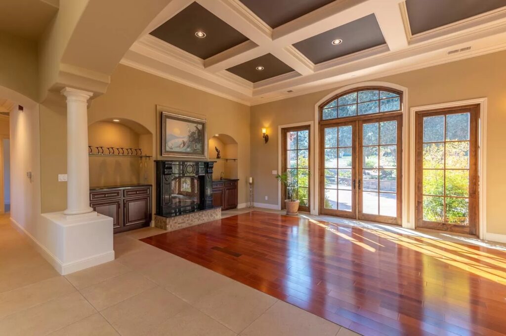 Spacious, sunlit living room with large windows, wooden floors, beige tile, coffered ceiling, and a black fireplace—expertly designed by a general contractor Pensacola for seamless flow from your next kitchen remodel or bath update.