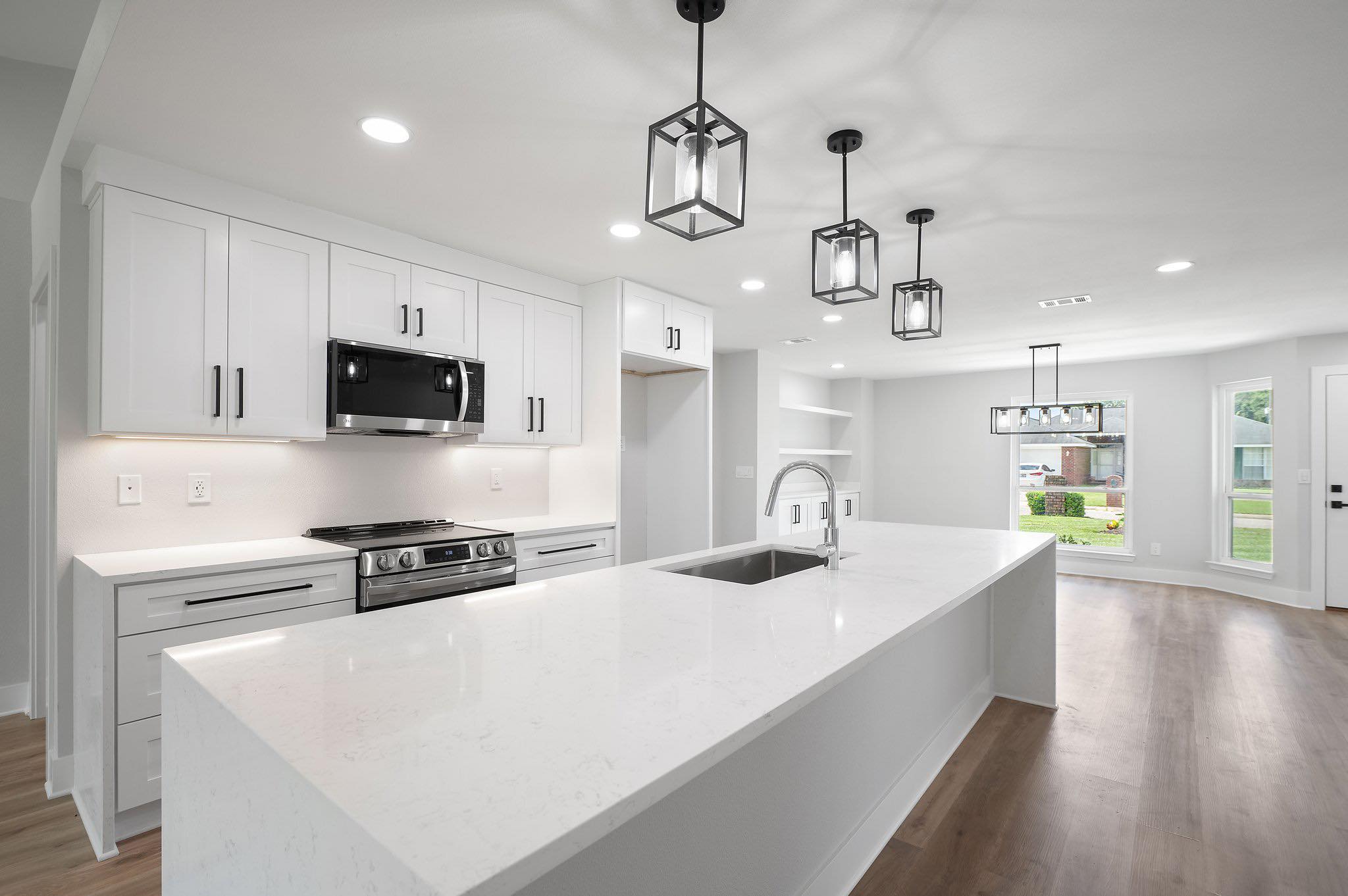 Modern white kitchen with a large marble island, built-in sink, stainless steel appliances, black pendant lights, wood floors, and bright natural light—perfect inspiration for your next kitchen remodel with a trusted general contractor in Pensacola.