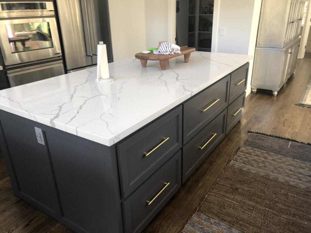A modern kitchen island with a white marble countertop, dark gray cabinets, and gold handles—crafted by an expert general contractor Pensacola. A paper towel holder and small wooden tray sit atop the counter, with wooden floors and rugs surrounding the island.