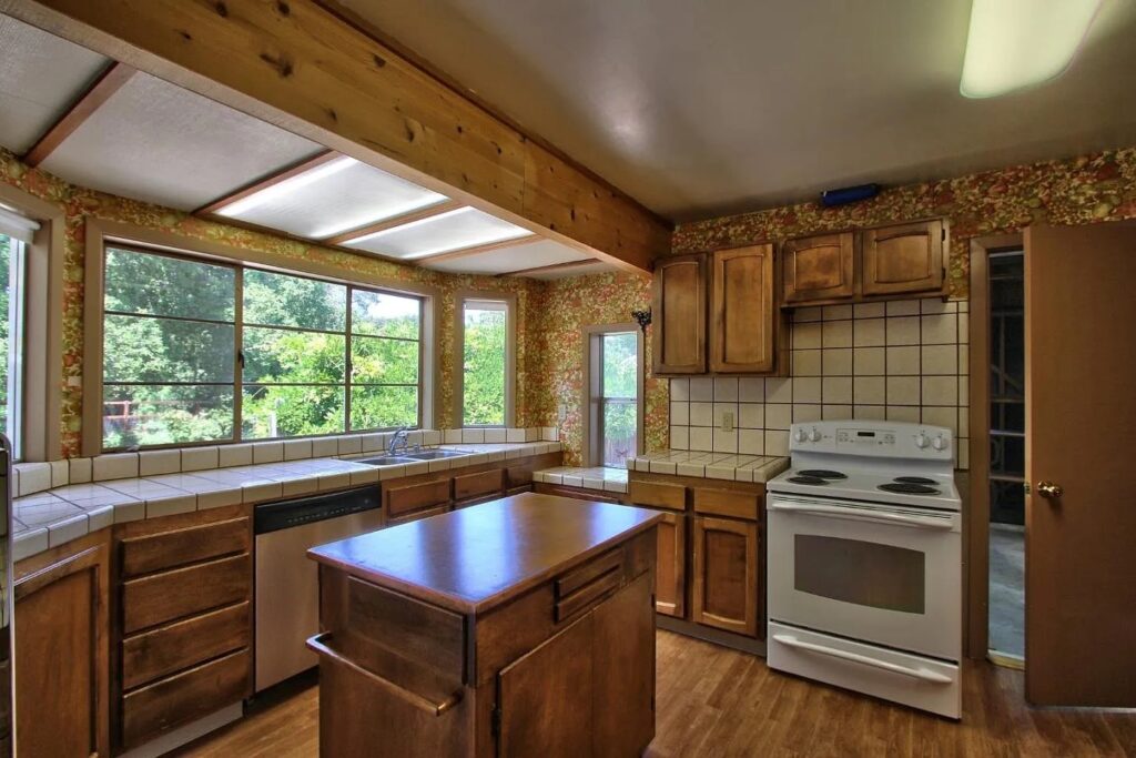 A kitchen with wooden cabinets, tiled countertops and backsplash, a white stove, dishwasher, small center island, large windows, and floral wallpaper; trees visible outside—perfect inspiration for your next Pensacola kitchen remodel.
