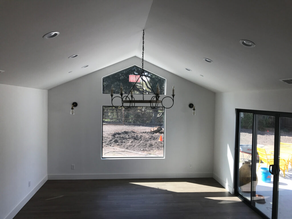 A modern, empty room with vaulted ceilings, a black chandelier, white walls, large window, and sliding glass doors to a patio with yellow chairs and dark wood flooring—perfect inspiration for your next Pensacola remodeling or home construction project. Sunlight streams in.