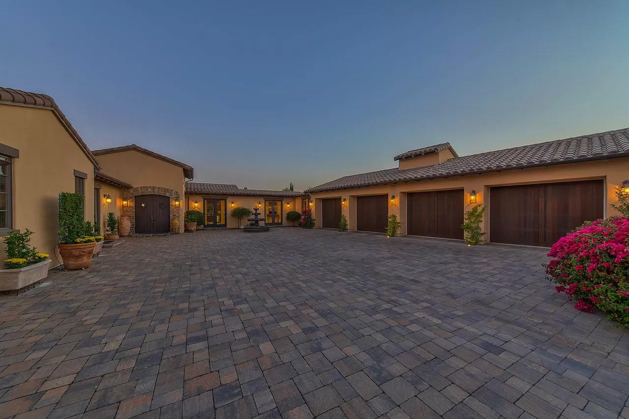 A spacious courtyard with stone pavers, surrounded by a large beige house—perfect for your next Pensacola remodeling project—features multiple wooden garage doors, warm exterior lights, potted plants, and vibrant flowering bushes at dusk.