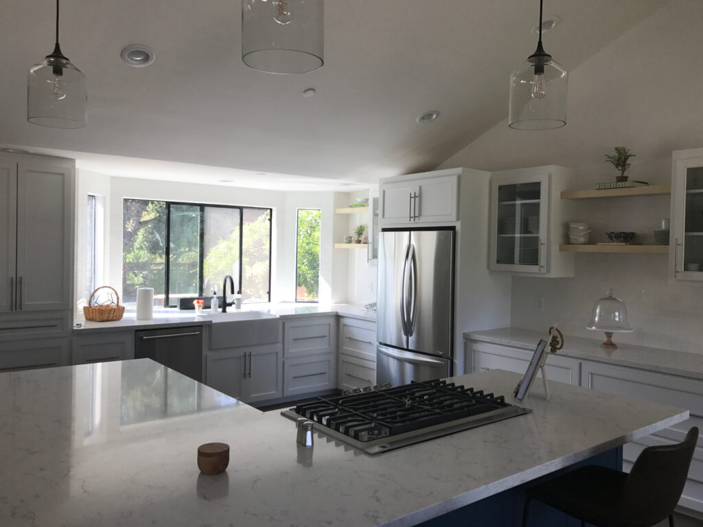 Bright modern kitchen with white cabinets, marble countertops, stainless steel appliances, large windows, open shelving, and a gas stovetop on a central island. Perfect for your Pensacola remodeling or kitchen remodel needs. Natural light fills the space.