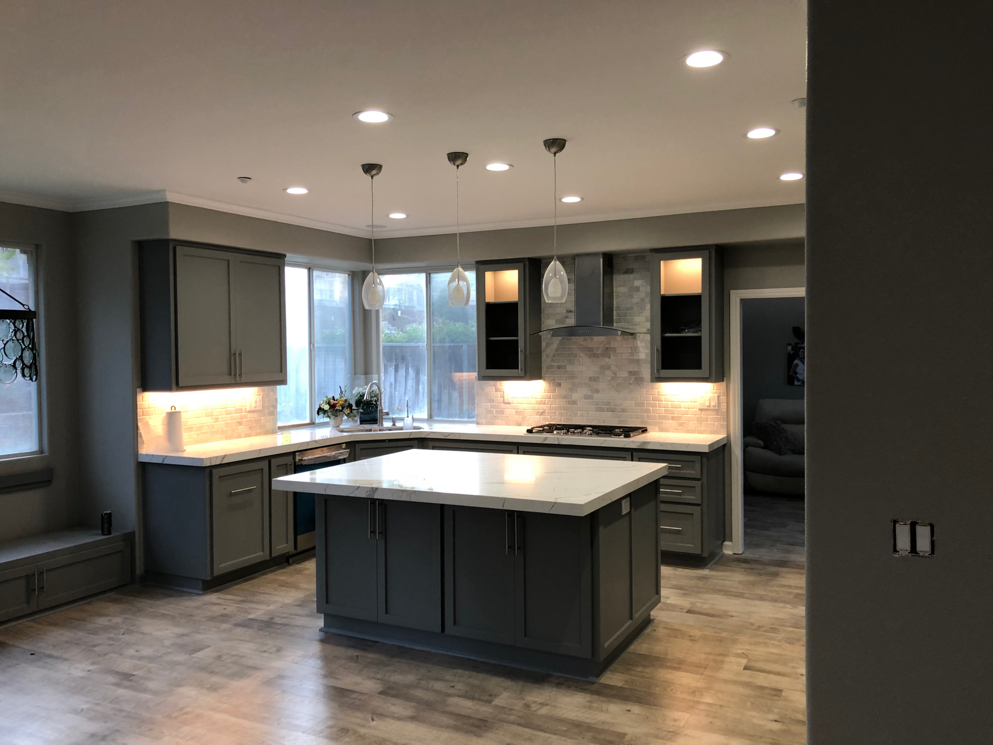 Modern kitchen with gray cabinets, white countertops, an island, pendant lights, stainless steel stove with hood, tile backsplash, wood floors, and large windows letting in natural light by a trusted general contractor Pensacola.