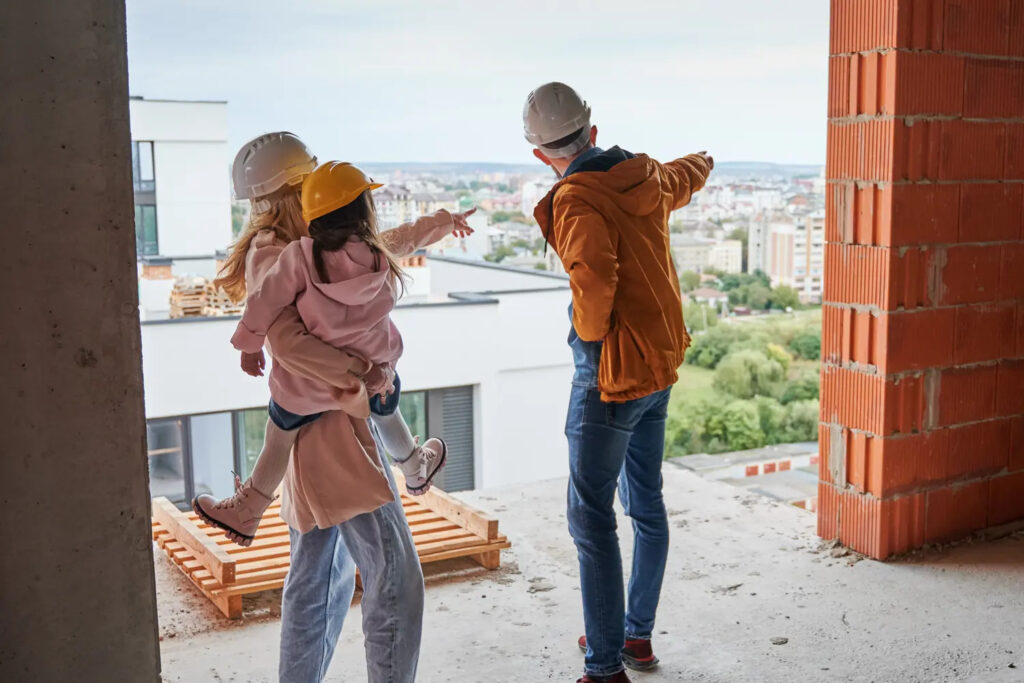A group of three people, including a child, all wearing hard hats, stand on an unfinished building floor and look out over a cityscape as one adult points towards the view, discussing ideas for their Pensacola remodeling project.