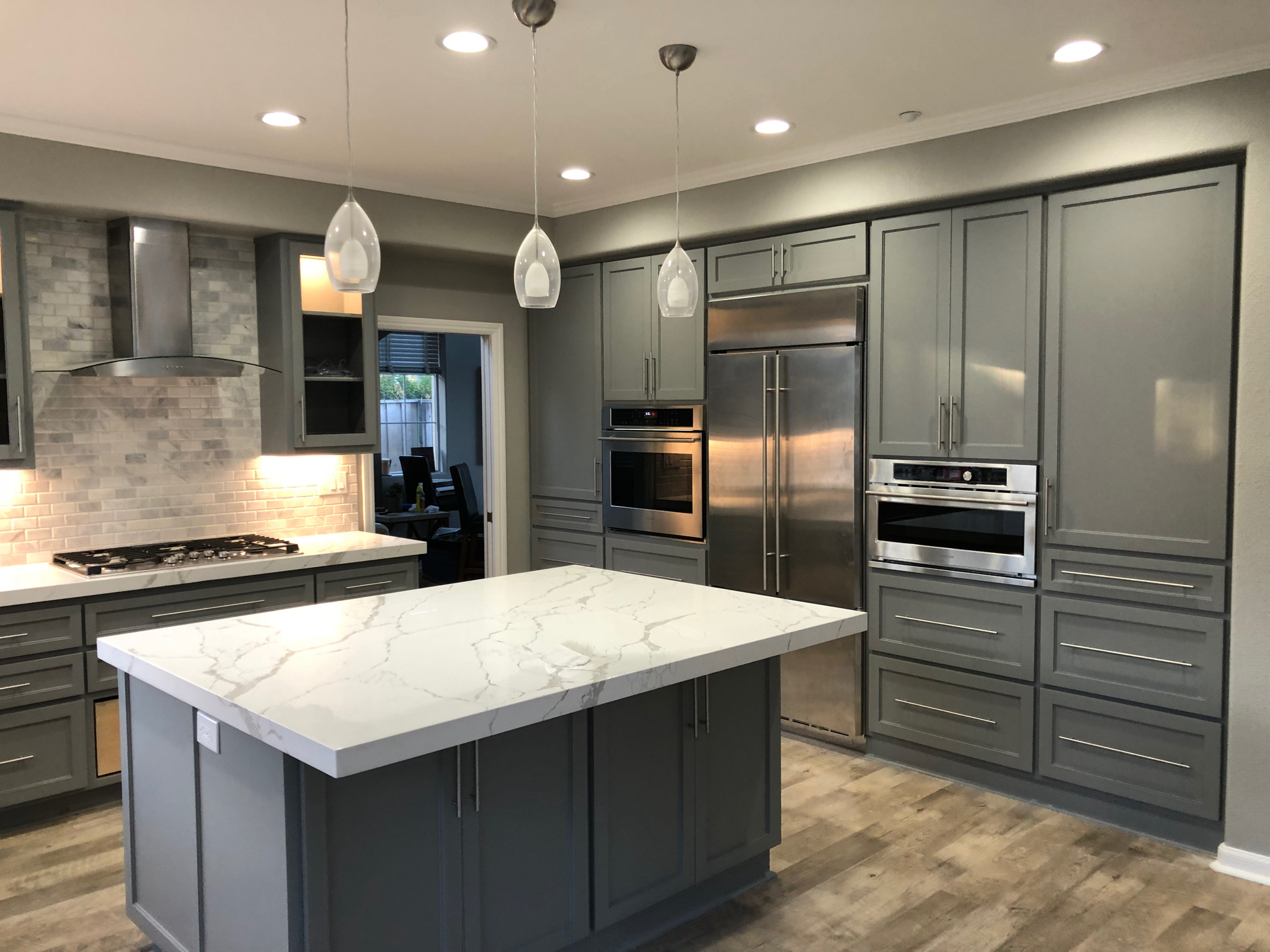 Modern kitchen remodel with gray cabinets, stainless steel appliances, a marble island countertop, pendant lights, and wood flooring. The backsplash is tiled in a light color, and recessed lights illuminate this Pensacola renovation.