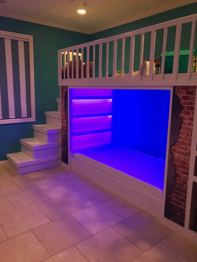 A modern, white bunk bed with stairs and built-in shelves is illuminated by blue LED lights. Perfect for a Pensacola remodeling project, the bed features a brick pattern on the sides, with teal walls and tiled flooring completing the look.