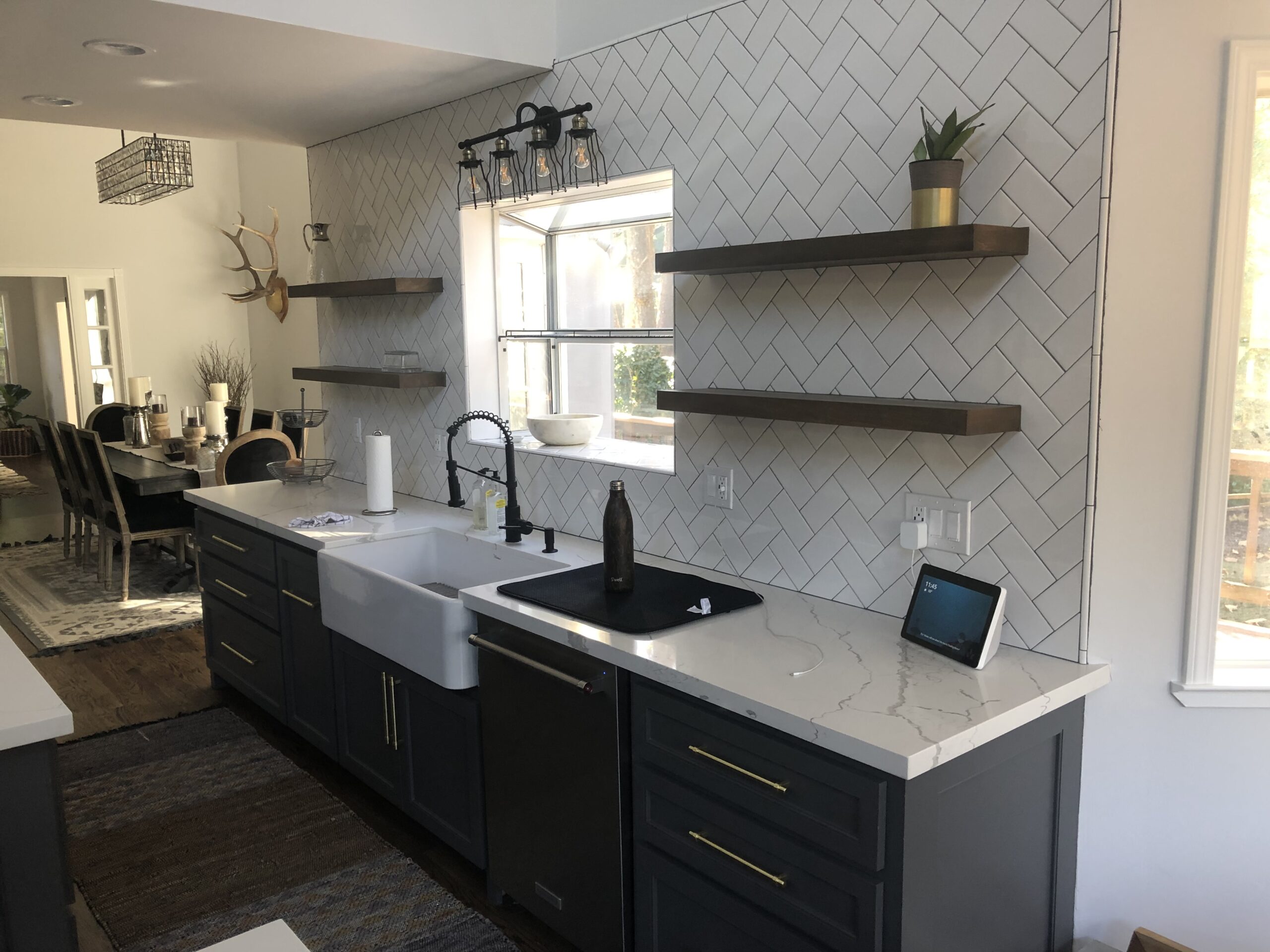 Modern kitchen with dark cabinets, white marble countertops, farmhouse sink, open wood shelves on a herringbone tile backsplash, potted plants, and natural light—crafted by a trusted general contractor Pensacola homeowners rely on.