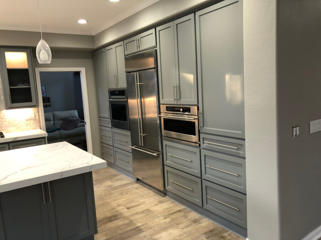Modern kitchen remodel with gray cabinets, a built-in stainless steel refrigerator, double wall ovens, and a marble-topped island. Light wood flooring and recessed lighting complete this clean, contemporary general contractor Pensacola design.