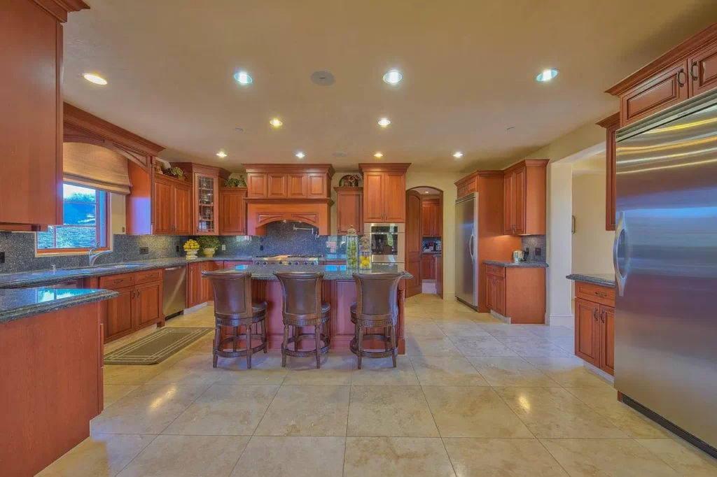 Spacious kitchen with wooden cabinets, a large central island with four chairs, granite countertops, stainless steel appliances, tiled floor, and recessed lighting. Perfect for your next Pensacola remodeling project or kitchen remodel.