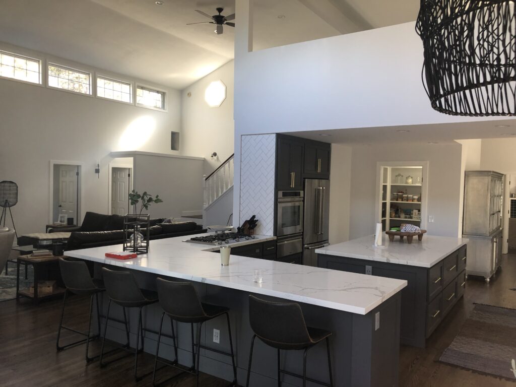 A modern open-concept kitchen remodel with a large white marble island, dark barstools, built-in ovens, and an adjacent living area. High ceilings and tall windows fill the space with natural light—perfect for your next Pensacola remodeling project.