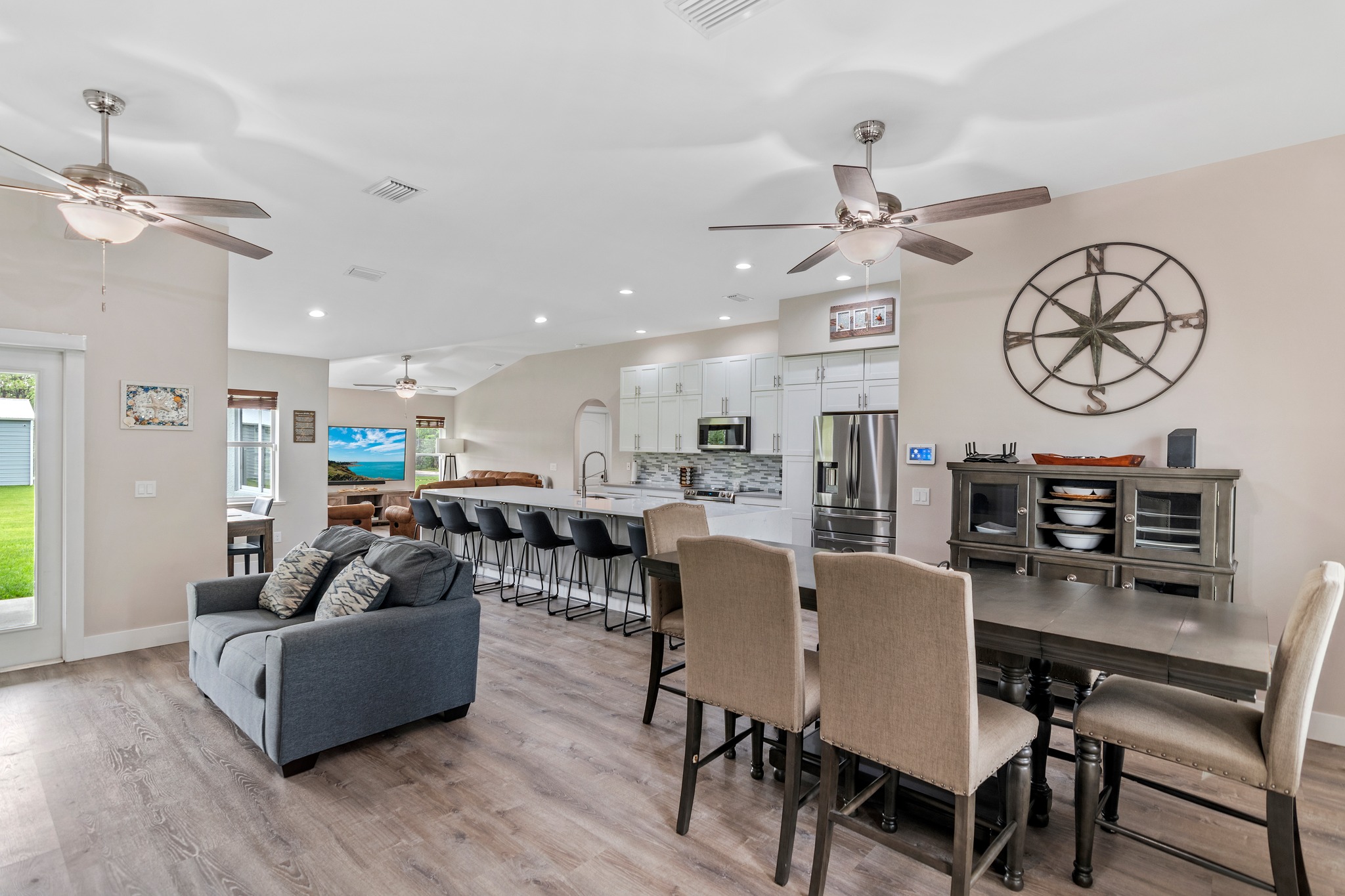 Open-concept living and kitchen area with gray sofa, dining table for six, modern kitchen remodel featuring bar stools, stainless steel appliances, and wood flooring. A large wall clock decorates the dining area. Trust a general contractor Pensacola for expert updates.