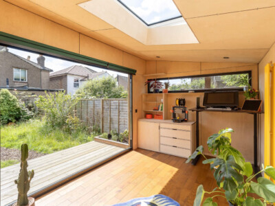 A bright garden office with large sliding glass doors, a skylight, wooden floors, a desk with a computer, shelves, and indoor plants—perfect inspiration for your next Pensacola remodeling or home construction project.