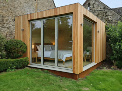 A modern wooden garden room with large glass sliding doors and windows reveals a cozy bedroom inside. Surrounded by green grass and neat bushes, it showcases the quality you’d expect from a top general contractor Pensacola trusts.