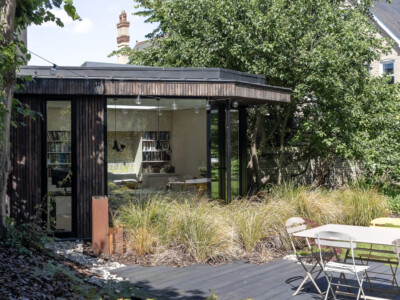 A modern garden office with large glass windows sits among trees and plants, designed by a general contractor Pensacola. A wooden deck and white outdoor table and chairs are in the foreground, while bookshelves are visible inside the office.
