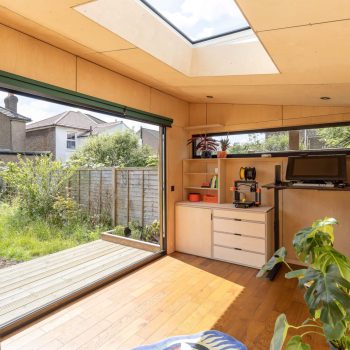 A bright garden office with large sliding glass doors, a skylight, wooden floors, a desk with a computer, shelves, and indoor plants—perfect inspiration for your next Pensacola remodeling or home construction project.