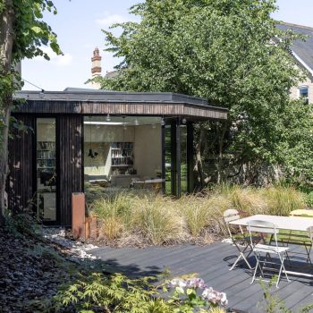 A modern garden office with large glass windows sits among trees and plants, designed by a general contractor Pensacola. A wooden deck and white outdoor table and chairs are in the foreground, while bookshelves are visible inside the office.